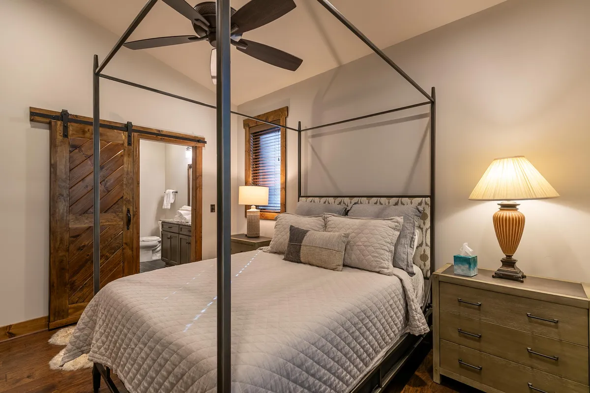 A cozy and calming primary suite featuring a queen canopy bed, a wooden sliding door, and a nightstand with a warm glowing lamp. The wood accents and gentle lighting create an inviting, restful ambiance perfect for relaxation after a day of adventure.