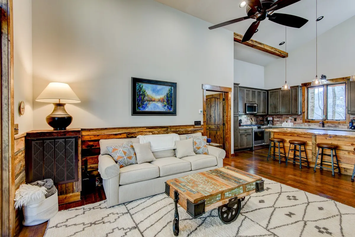 The image shows a bright, open-plan living and kitchen area, with a plush sofa, coffee table, visible kitchen space, and artful touches. Natural light fills the room, highlighting the seamless transition between spaces for relaxation and entertaining.