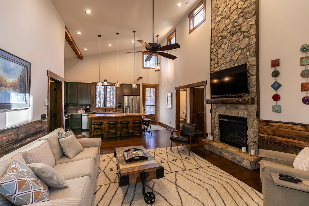 This image displays an open-concept living area featuring a comfortable sofa oriented toward a rustic stone fireplace. The kitchen island is visible in the background, offering a central gathering spot. The warm wood tones and inviting layout create a seamless flow between relaxing and social spaces.