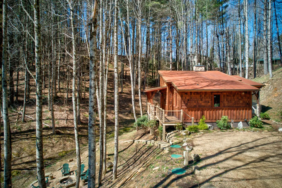 This exterior image shows a cozy wooden cabin nestled among trees, with a visible fire pit area and seating where guests can gather. The deck, surrounded by natural beauty, is perfect for unwinding after a day of adventure.