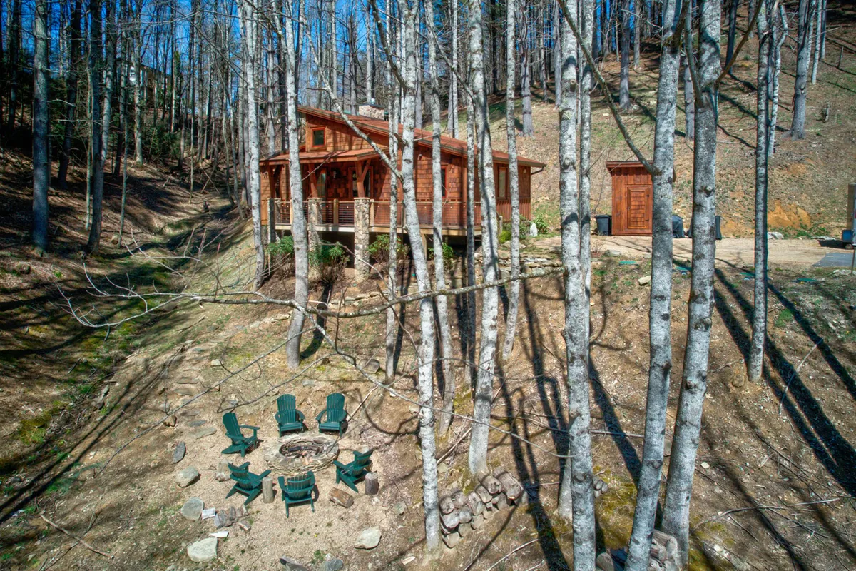 A cozy cabin sits among tall trees with a prominent fire pit area in the foreground, encircled by seating. The inviting setup suggests gatherings around a crackling fire, with space for marshmallow roasting or quiet relaxation under the stars.
