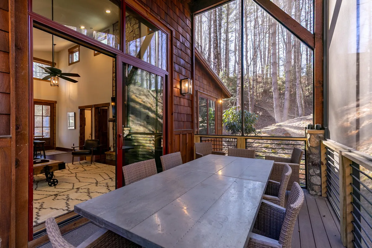 This image features a wooden outdoor dining table positioned on a patio or deck, surrounded by large windows and enveloped by lush trees. The scene conveys a comfortable, welcoming atmosphere ideal for enjoying meals outdoors or relaxing with a morning coffee while immersed in nature.