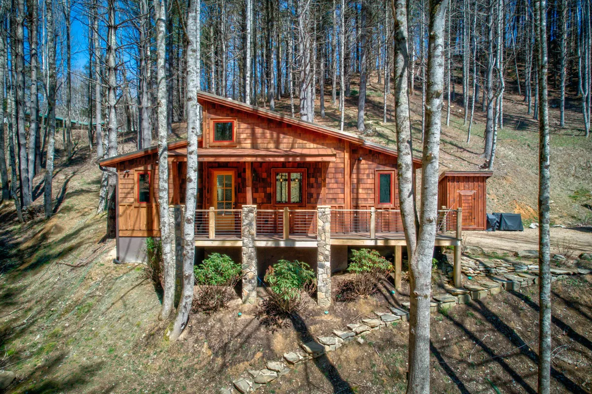 The image features the warm wooden exterior of Doe Run Cottage nestled among tall trees, with a spacious deck supported by impressive stone pillars. The inviting deck is perfectly positioned for watching sunsets, sipping morning coffee, or gathering around a fire pit in the evenings, all surrounded by the peaceful beauty of the North Carolina mountains.