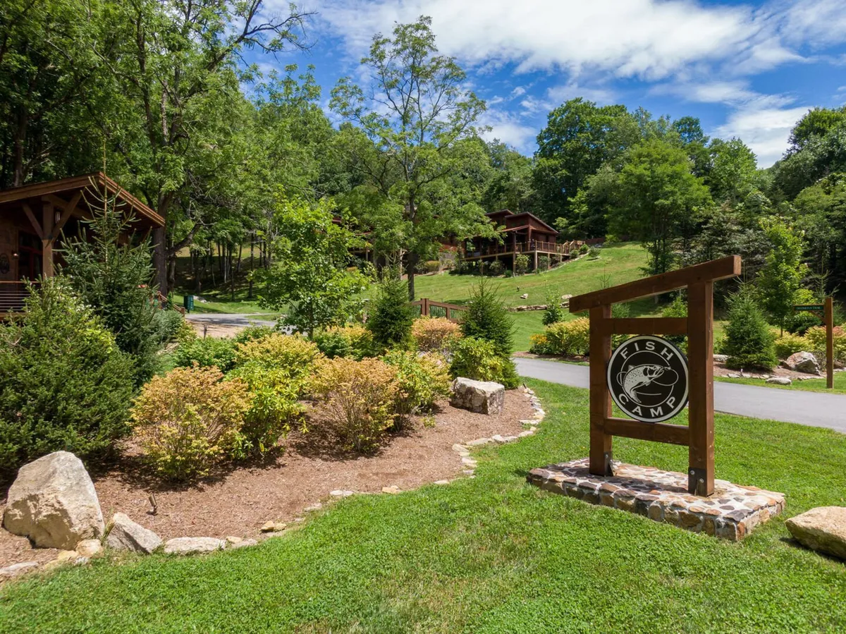 An exterior view featuring the Fish Camp sign, beautifully landscaped gardens, and cozy cabins in the background. The scene evokes peaceful adventure, with the exclusive fishing area highlighted and the sense of a private, nature-focused retreat.
