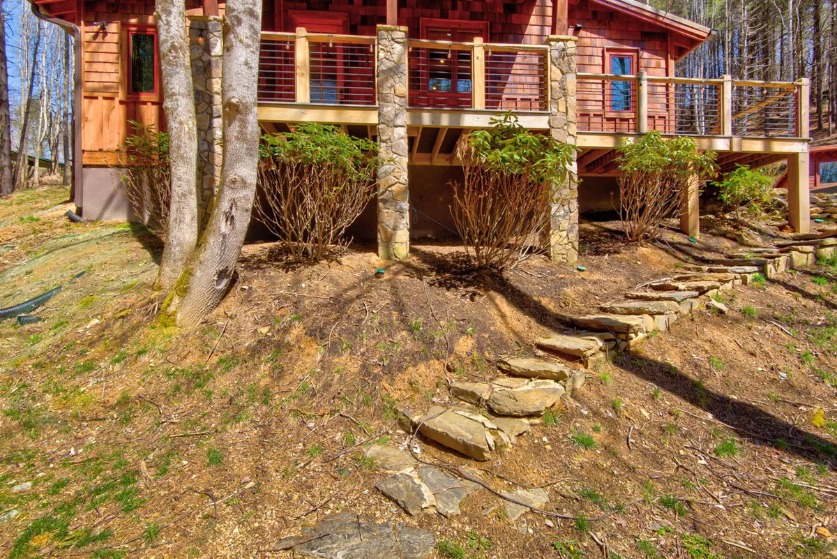 A wooden deck overlooks a landscaped area and connects to a winding stone pathway, all surrounded by mature trees. The scene suggests a tranquil transition between the cottage and the natural world, inviting guests to explore or relax just steps outside.