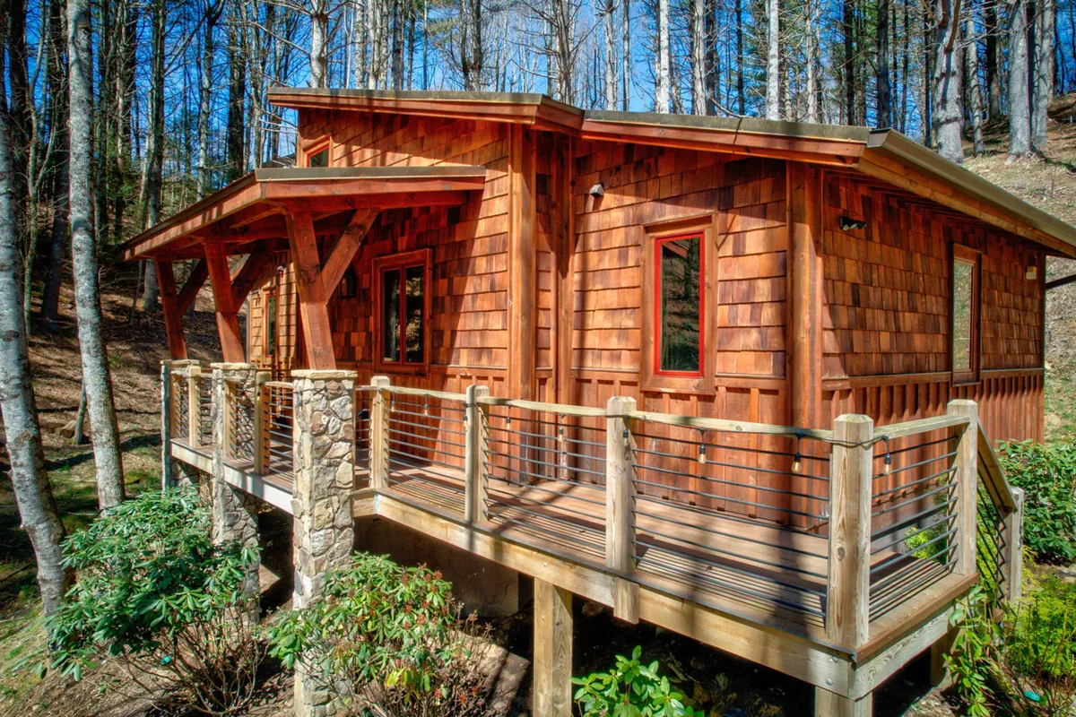 This exterior image highlights the wooden siding, sturdy stone pillars, and a deck railing, all surrounded by lush greenery. The deck is a focal point for relaxation, with ample space for guests to unwind while enjoying the tranquil mountain setting. The soft evening light evokes a sense of peaceful retreat, perfect for sipping wine or gathering with friends.