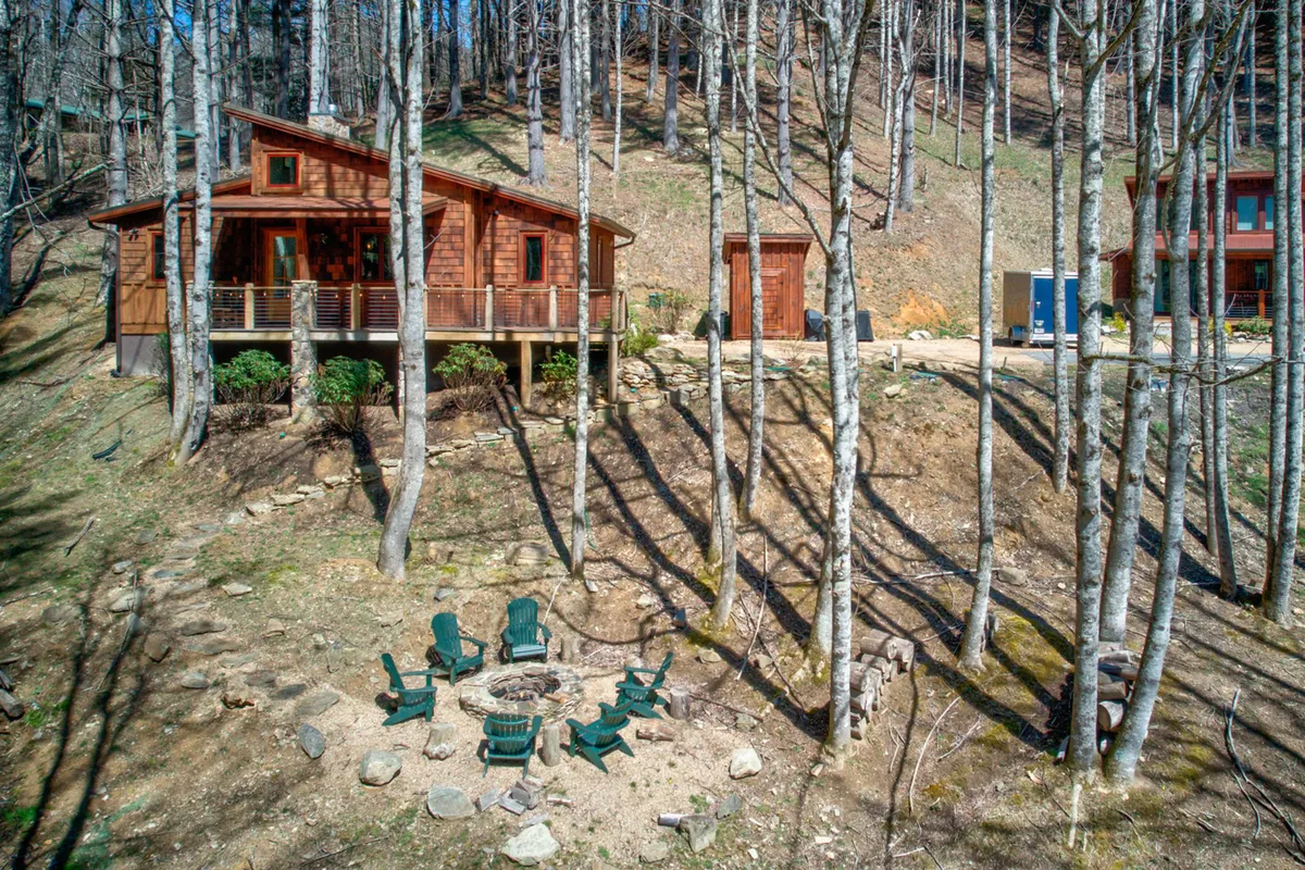 This image captures a charming wooden cabin nestled among trees, with a prominent fire pit area in the foreground and a tranquil deck for relaxing. The lush greenery surrounding the cabin highlights the connection to nature and provides an inviting space for outdoor activities with pets.