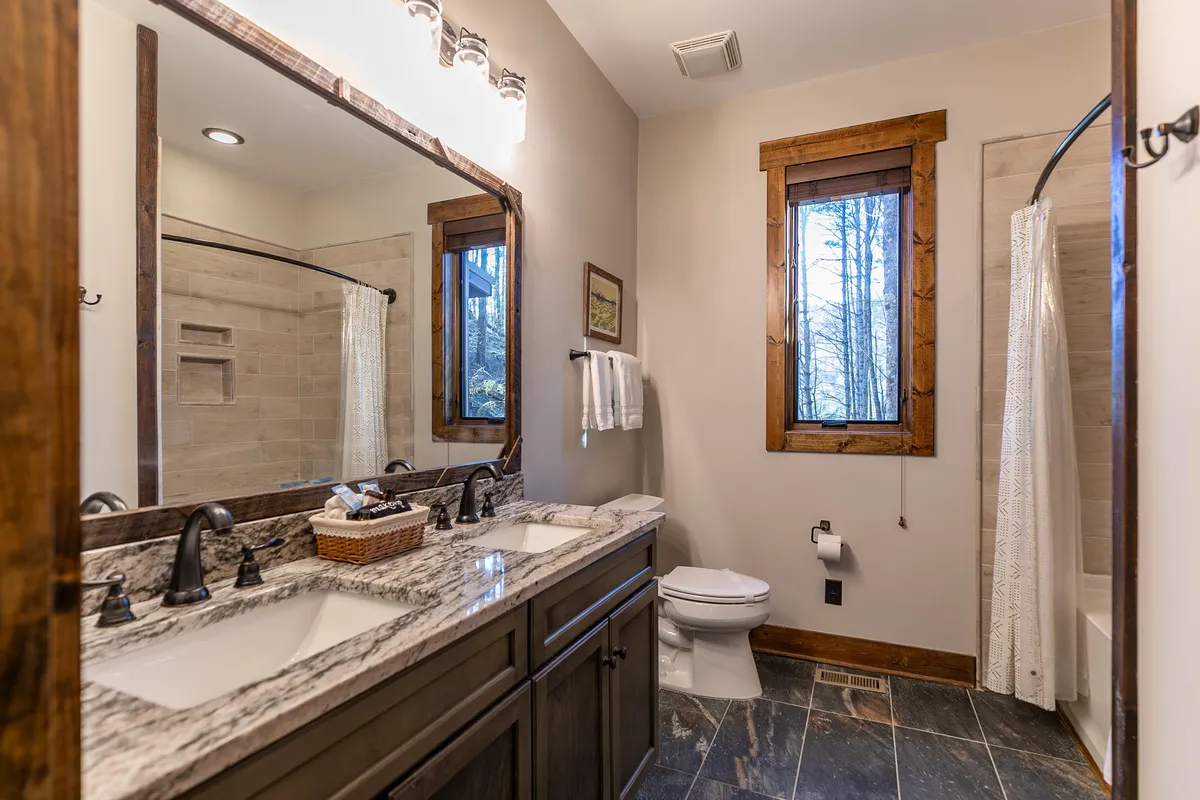 A bright, refreshing bathroom featuring a double vanity with sinks, a large mirror, a window letting in natural light, and a visible shower area. The space feels calm and rejuvenating, ideal for winding down after a long day.